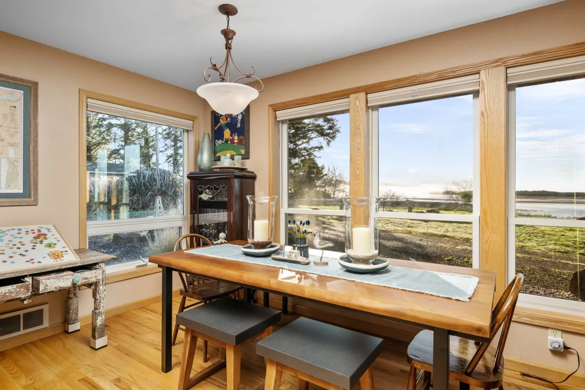 A welcoming dining area anchored by a large wooden table and surrounded by oversized windows. Elegant decor, a decorative cabinet, and artwork create an inviting atmosphere, while the expansive windows offer mesmerizing views of the water, ideal for shared meals and meaningful conversations.