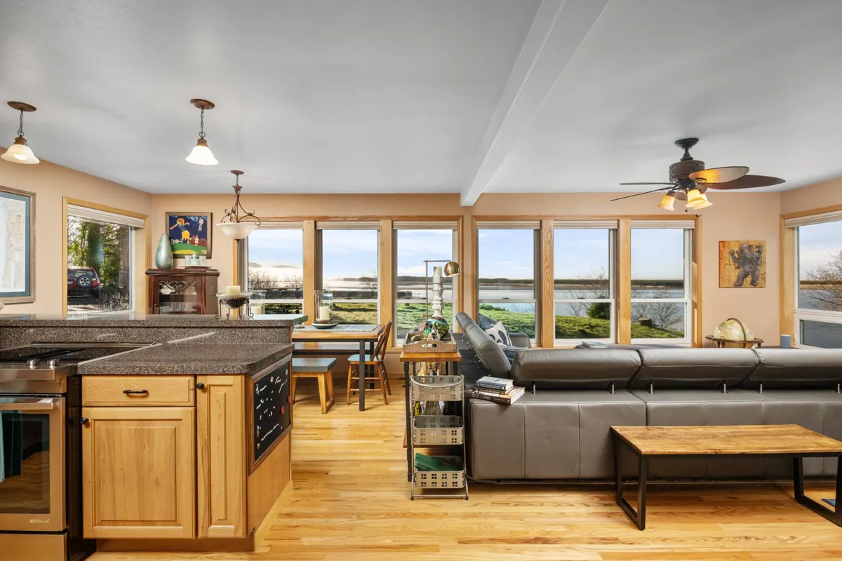 A bright, open-concept living area featuring large windows that frame expansive water views. The image captures the seamless flow between the kitchen island, comfortable seating area, and dining table, all bathed in natural light. The panoramic windows bring the outside in, emphasizing the home's connection to its stunning coastal setting.