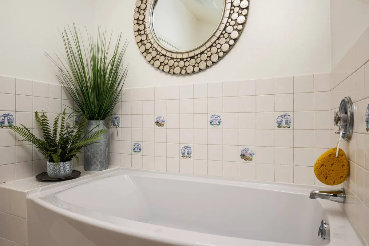 A peaceful bathroom scene featuring a bathtub, decorative mirror, a houseplant, and soothing wall tiles, hinting at a tranquil space for relaxation and self-care.