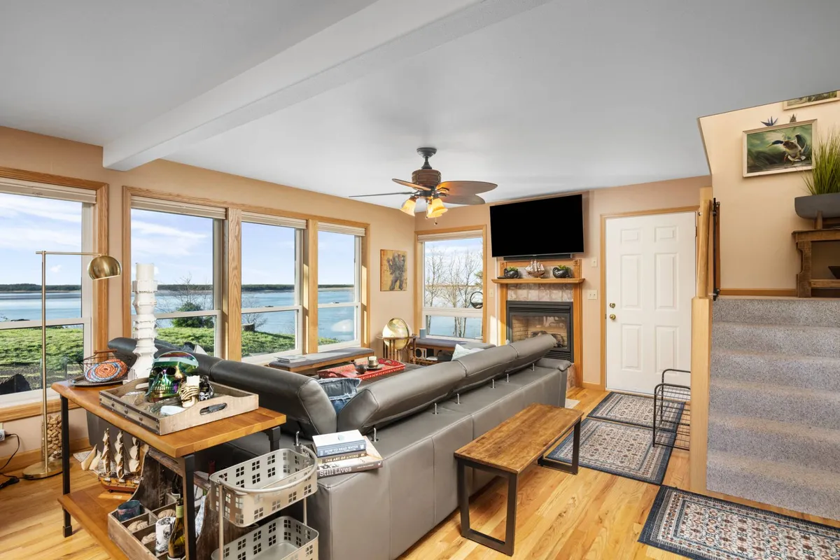 The spacious living room features a comfortable sofa, a fireplace, and large windows overlooking the water. Shelves with decorative items add personality and warmth. The setup is perfect for relaxing with loved ones, watching the sunset, or enjoying a cozy evening by the fire.