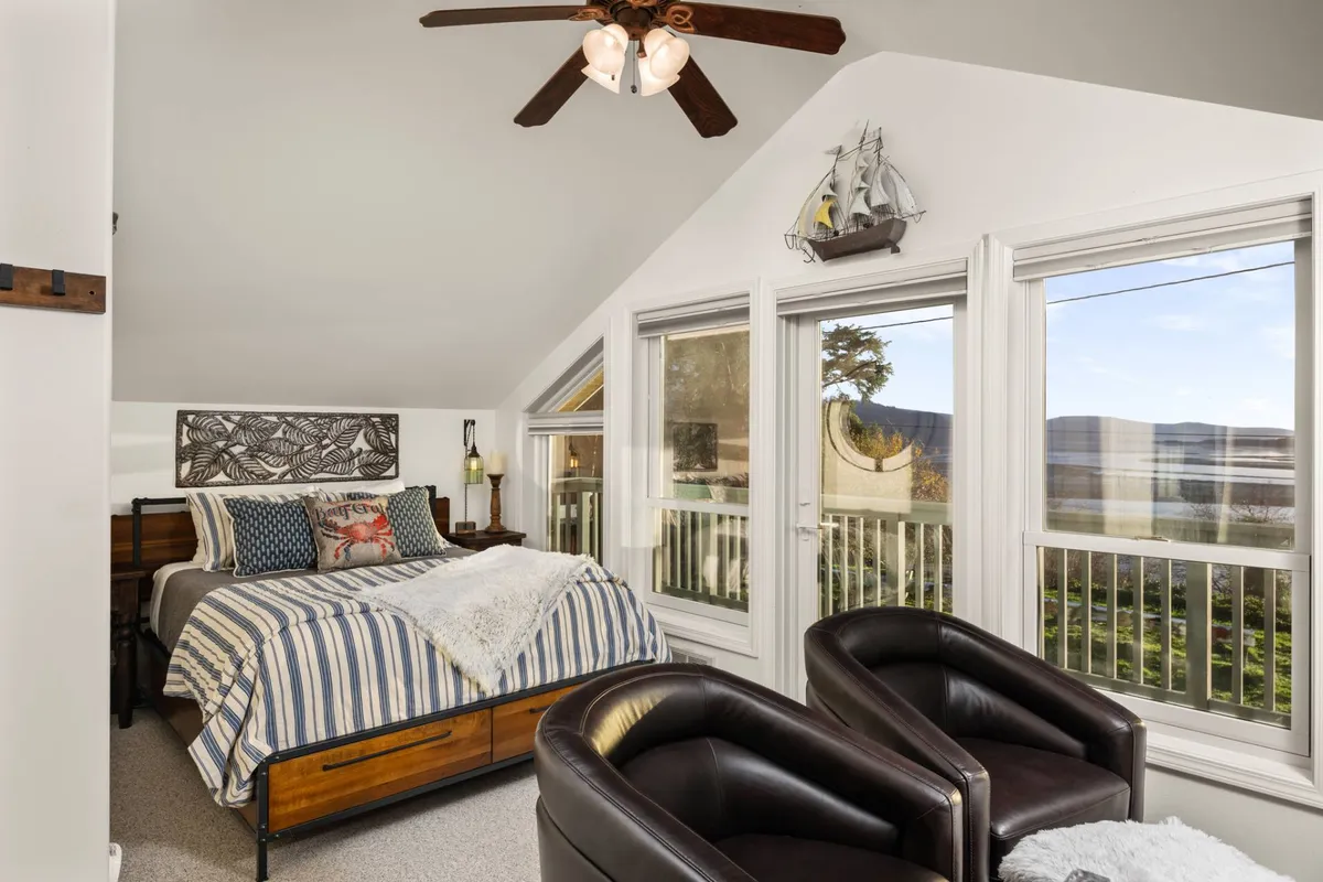 A serene upstairs bedroom with a queen bed adorned in striped bedding, large windows offering a tranquil view, and two leather chairs set up for relaxing or reading by natural light.