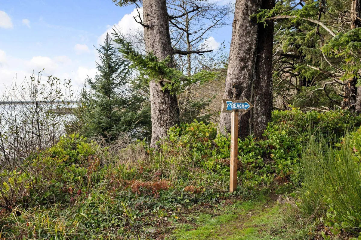 A rustic wooden sign points the way to the beach, surrounded by tall trees and lush coastal greenery. The path beneath is dappled with sunlight, inviting visitors to embark on a tranquil nature walk through the nearby forest trails, hinting at the proximity of exploration and beach adventures.