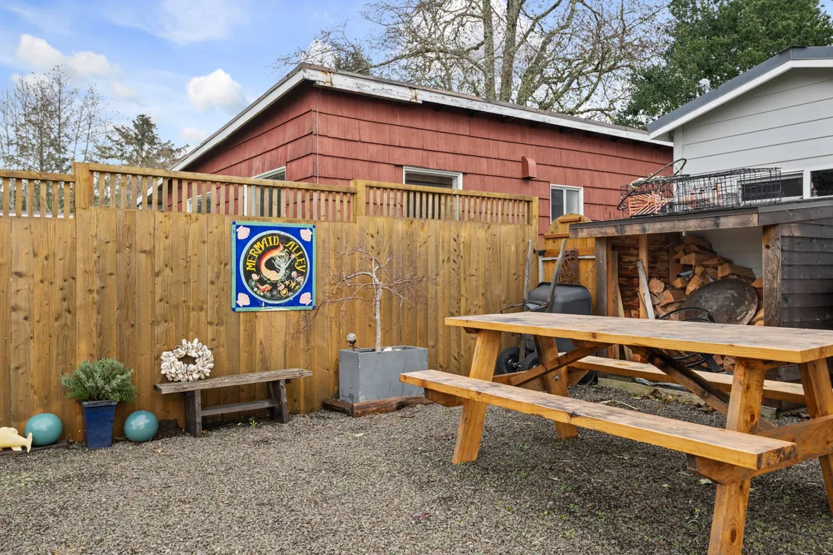 This image shows a casual and friendly outdoor setting featuring a wooden picnic table, decorative wall art, a stack of firewood, and lush potted greenery. The area is clearly designed for social gatherings, relaxed meals, and evening conversations by the fire pit.